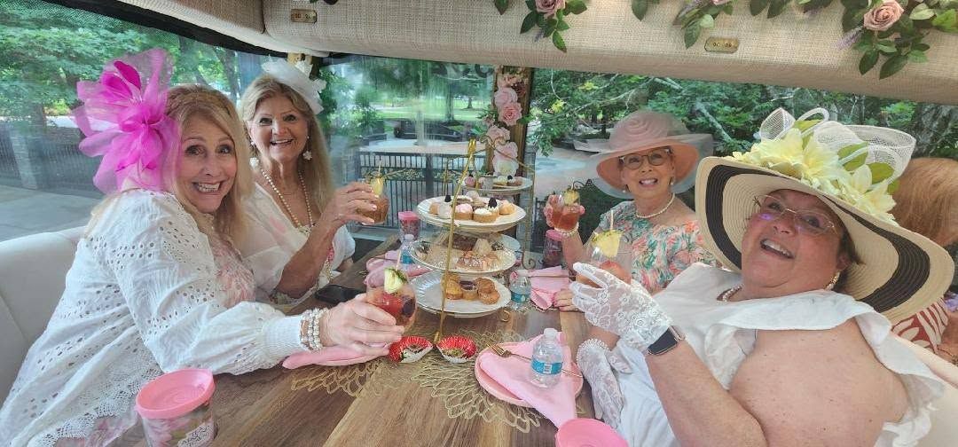 Four friends in pastel hats enjoy tea and pastries at a garden table, smiling outdoors.