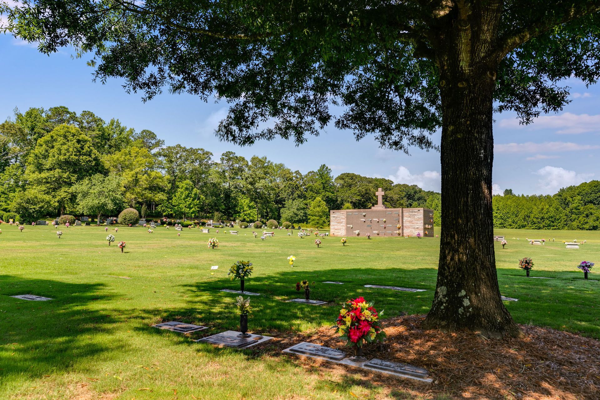 Tree-shaded cemetery lawn with headstones, a flowered grave, and a stone mausoleum in the distance