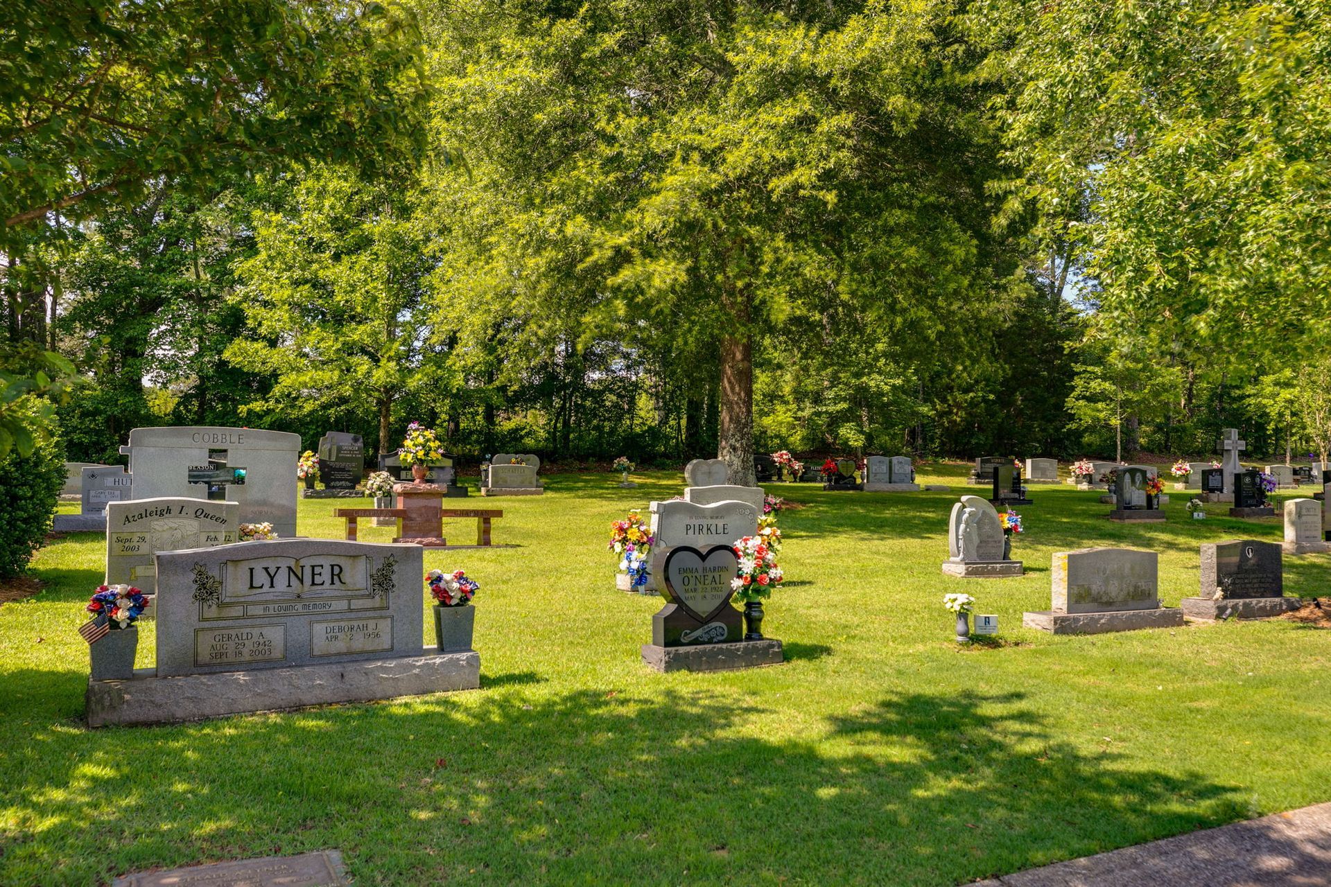 Gravestones in a sunny cemetery beneath large green trees, with flower arrangements on several markers