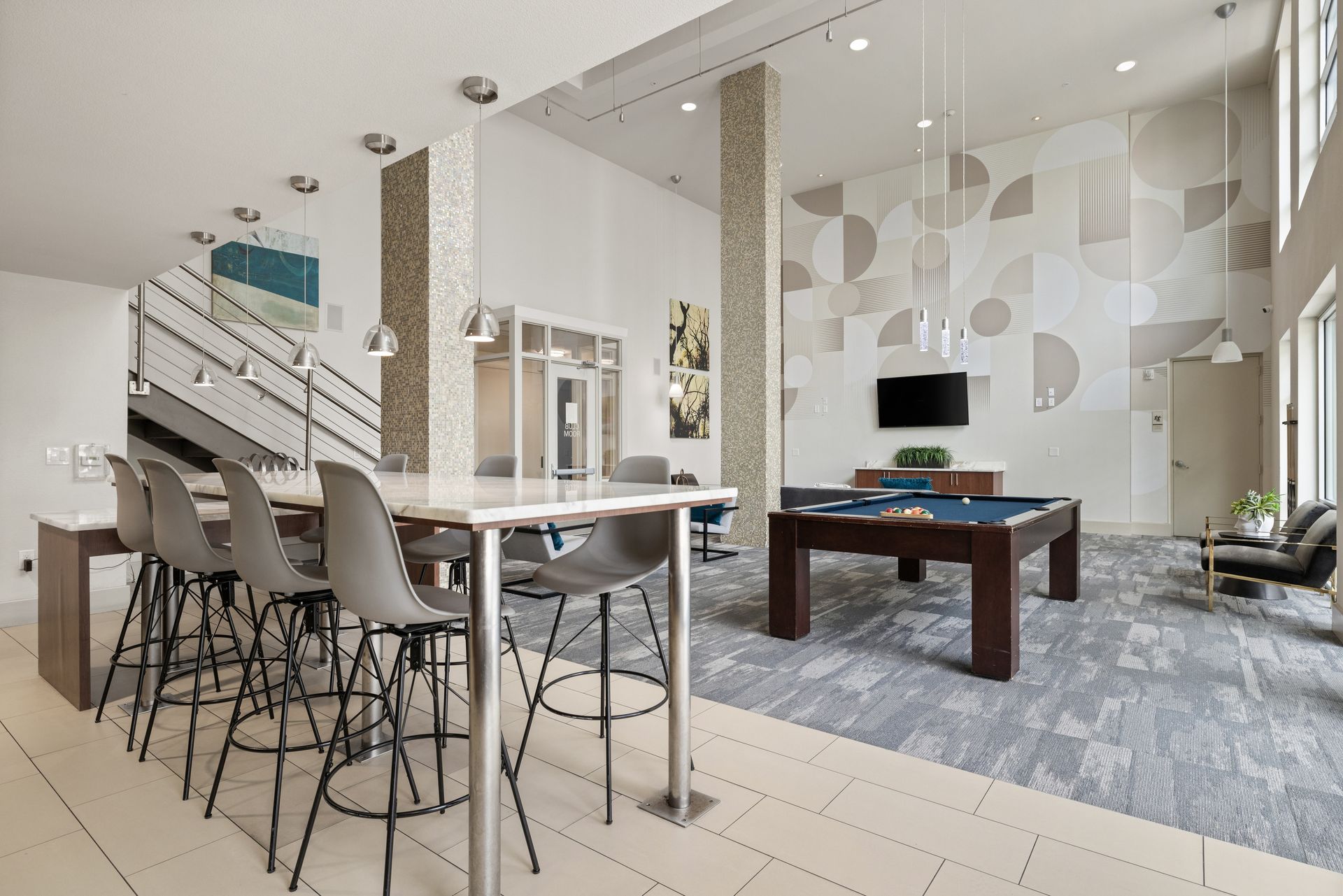 Modern open-concept clubhouse with gray furniture, bar stools, staircase, and wall-mounted TV at Sabina in Austin, TX.