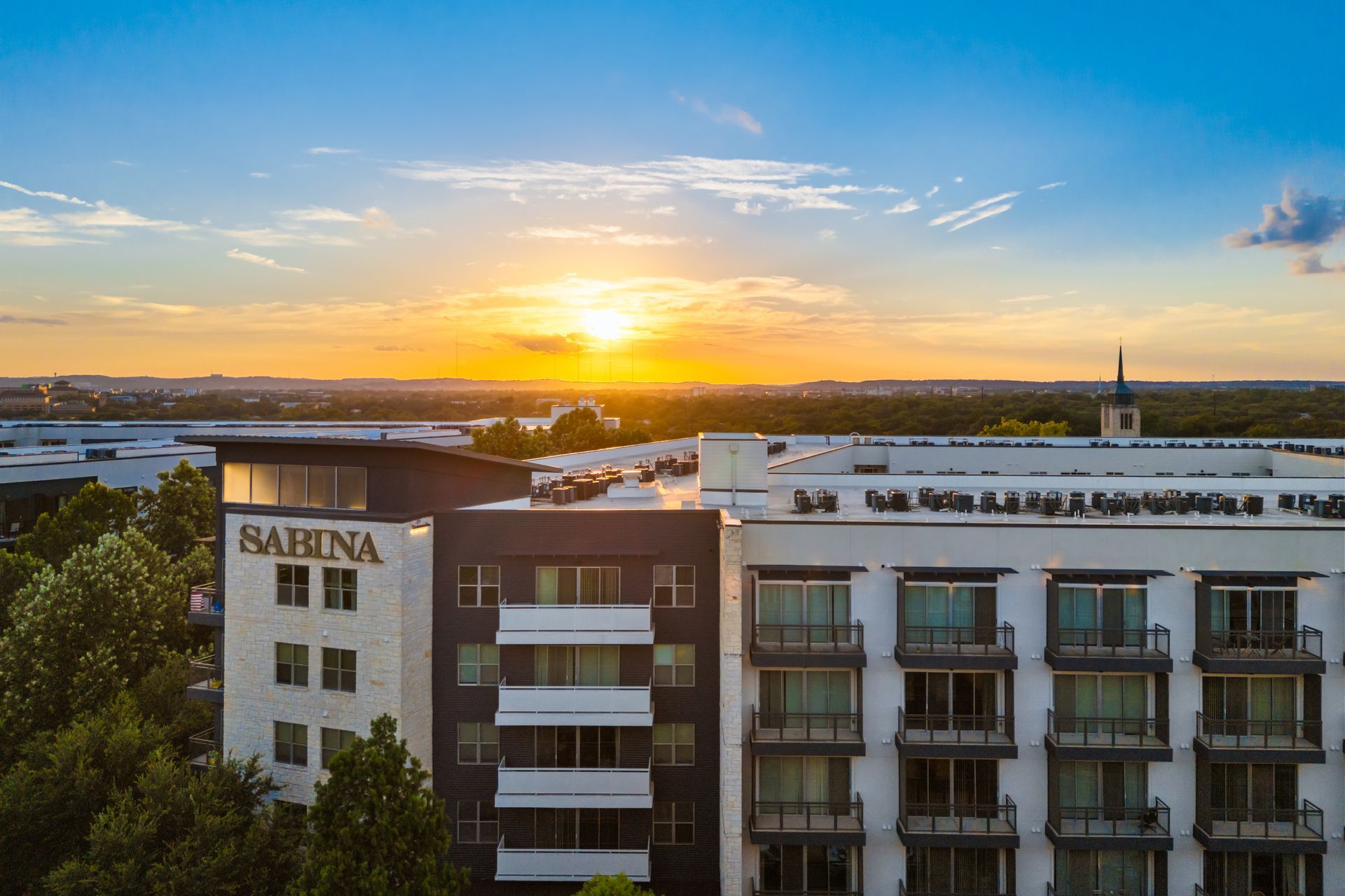 Sunset over a city skyline with a river and modern buildings, one labeled “SABINA”