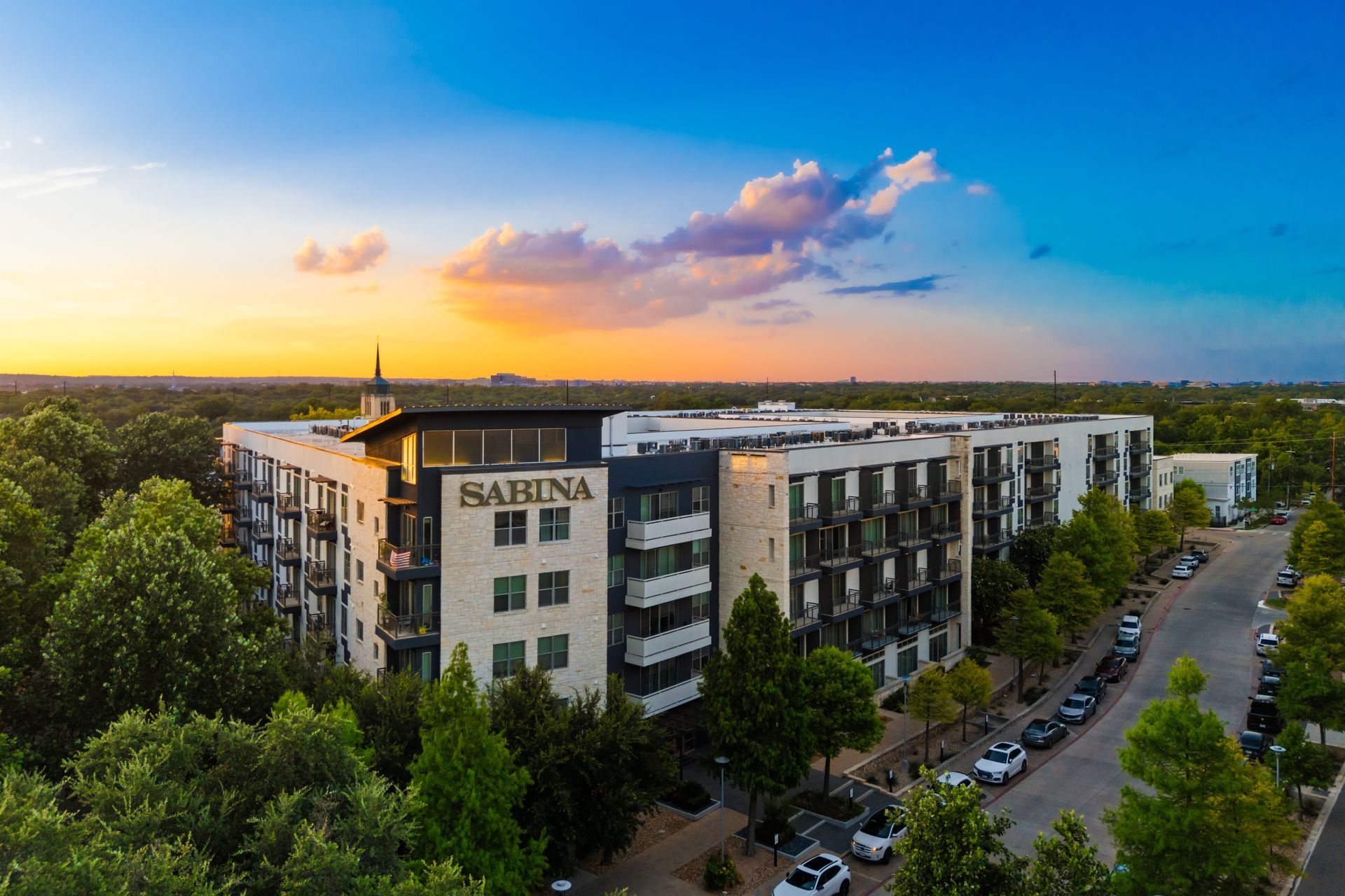 Aerial view of a modern apartment building at sunset with trees and parked cars along the street