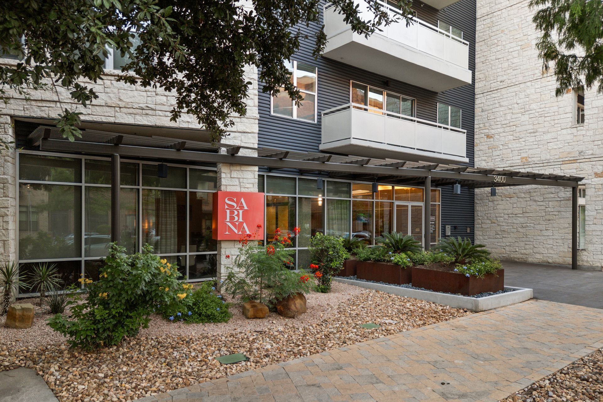 Apartment building entrance with glass doors, landscaped rocks and shrubs, and a red banner by the walkway