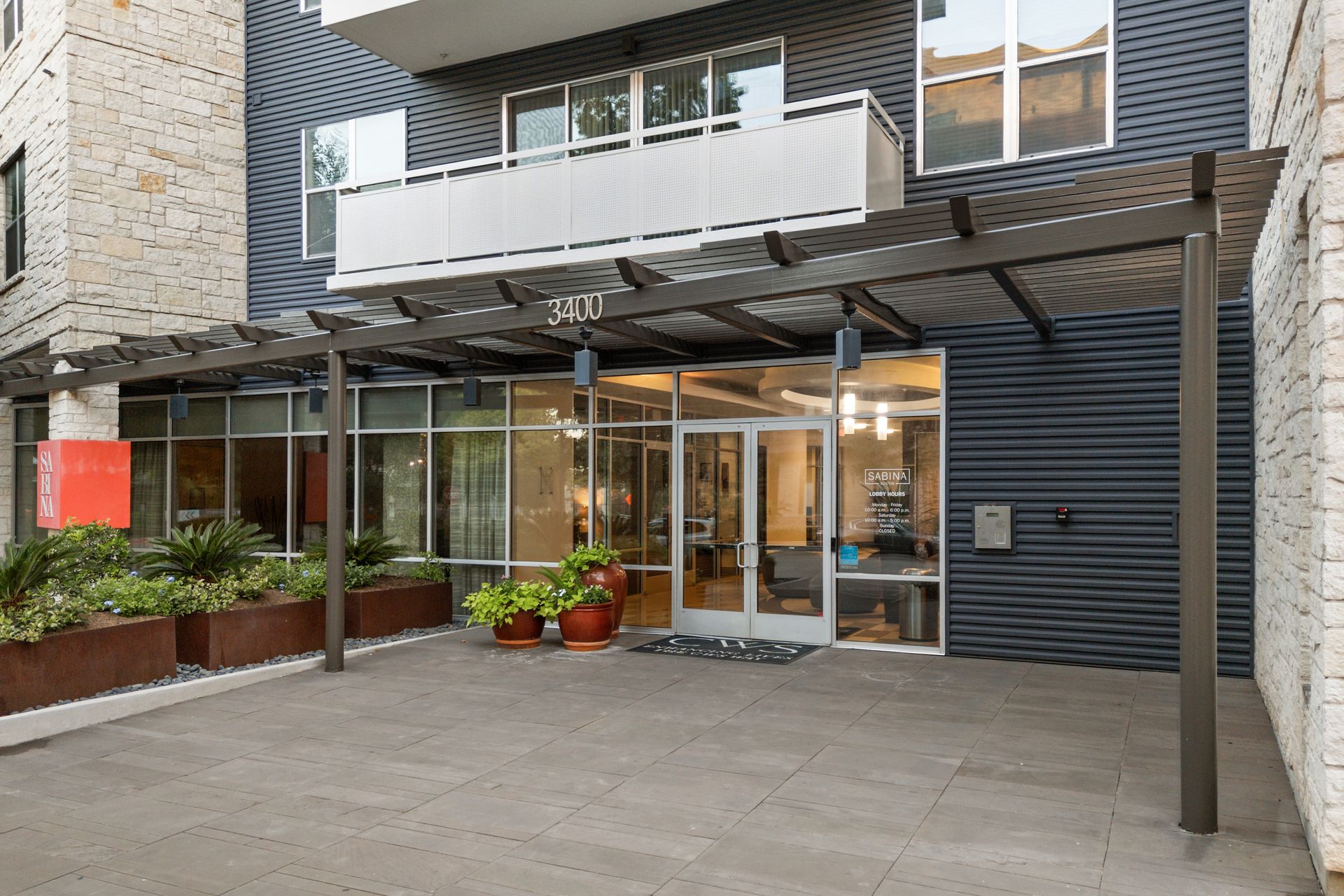 Modern apartment building entrance with glass doors, balcony above, and potted plants by the walkway