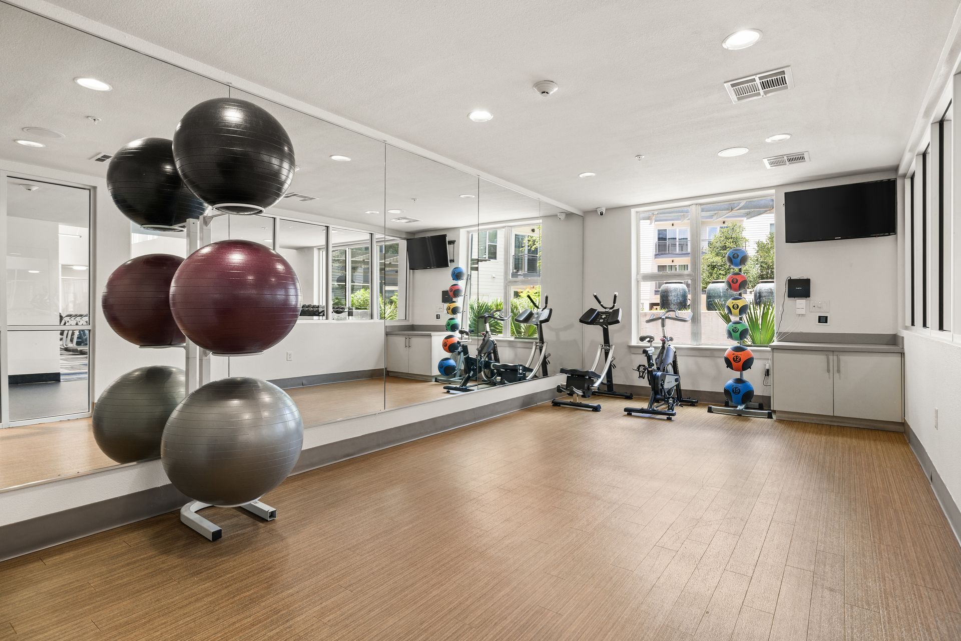 Bright gym studio with exercise balls, mirrors, wood floor, and stationary bikes by the windows.