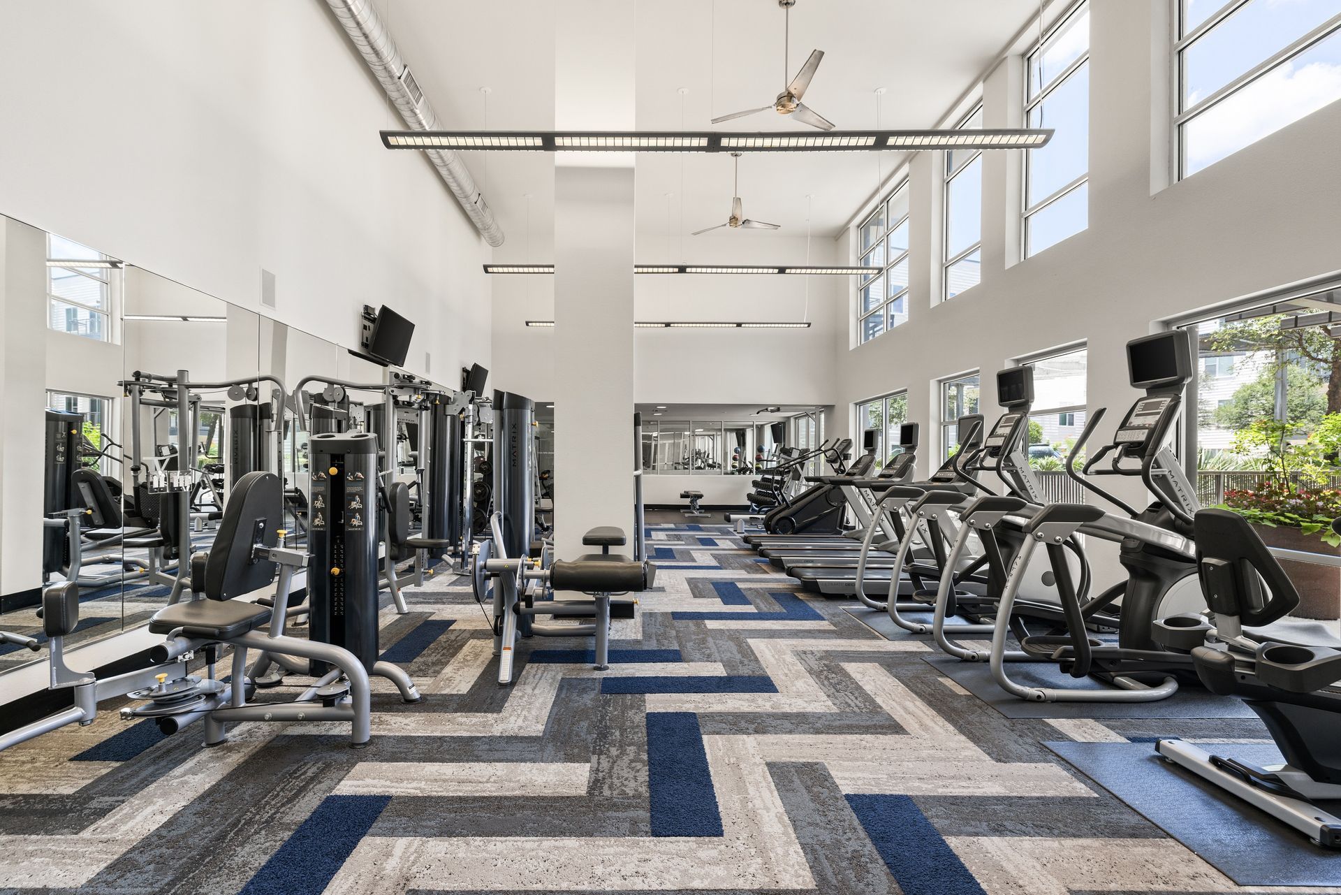 Modern gym with rows of cardio and weight machines, white walls, and blue-gray patterned flooring.