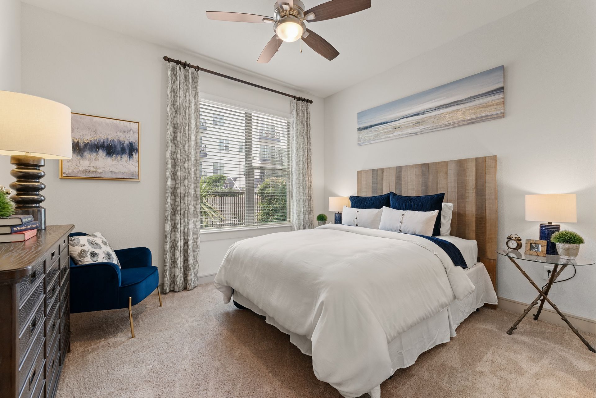 Bright bedroom with a bed, blue chair, lamps, window, and ceiling fan in neutral tones