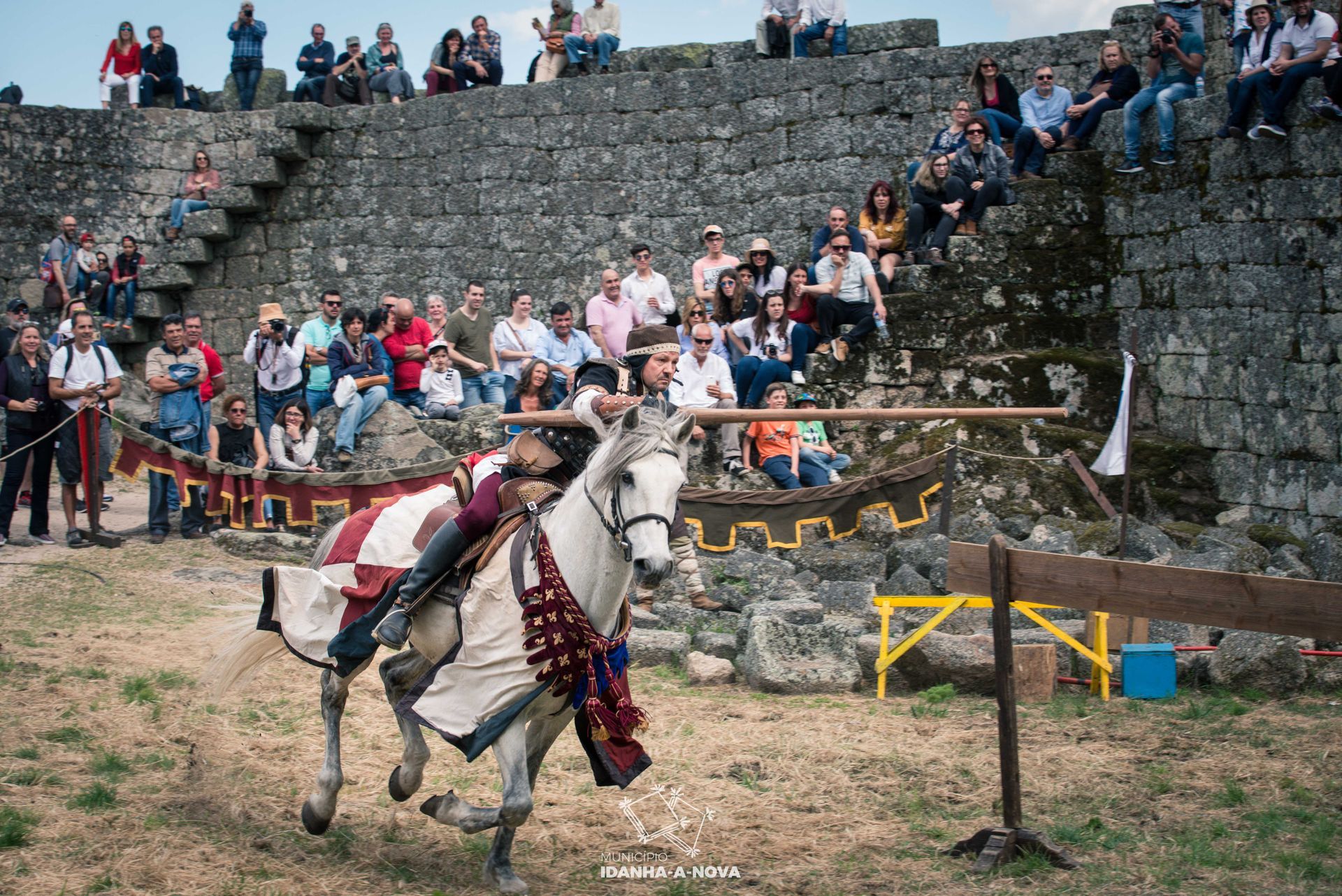 Cavaleiro montado num cavalo branco, com lança numa arena medieval e espectadores nas muralhas de pedra.