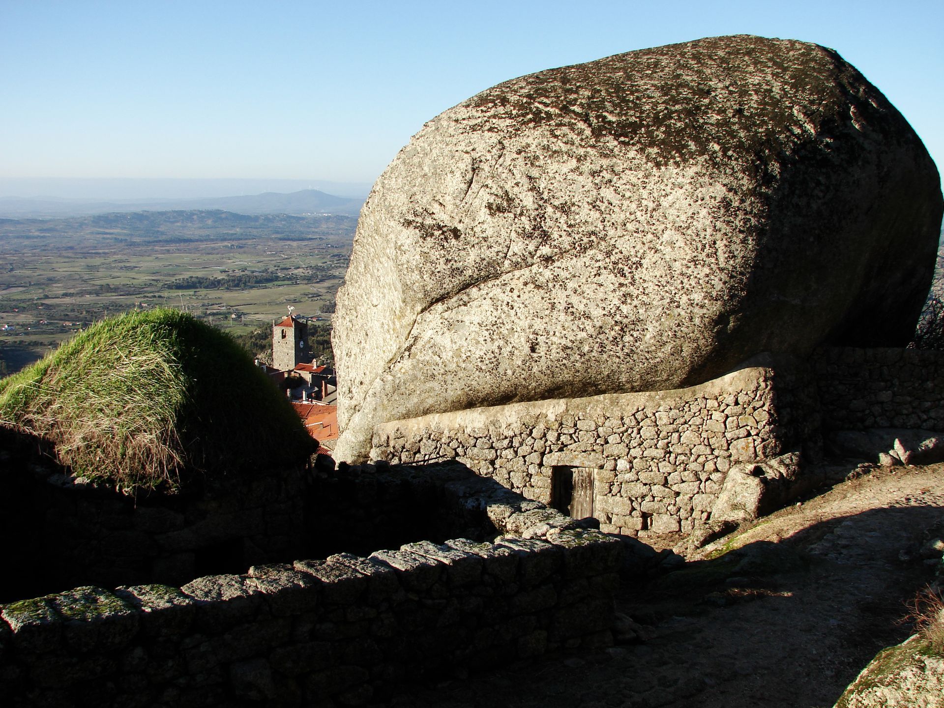Grande bloco de granito com vista para um vale, com um muro de pedra em primeiro plano e uma paisagem distante ao fundo