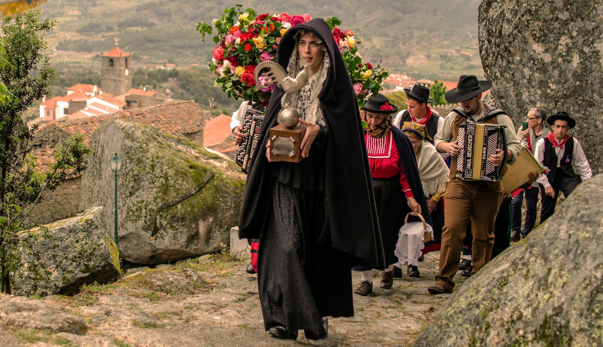 Mulher vestida de preto sobe a encosta com flores, seguida por músicos numa procissão rural pela colina