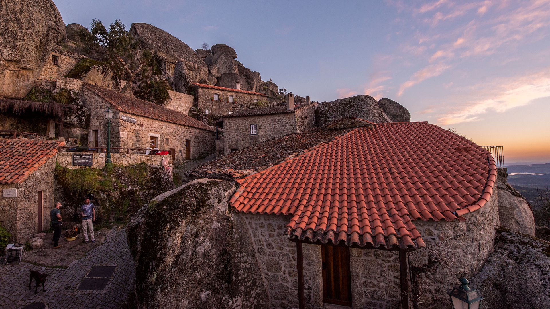 Casas de pedra numa aldeia, com telhados de telha vermelha, numa encosta rochosa ao pôr do sol, com vista para o mar.
