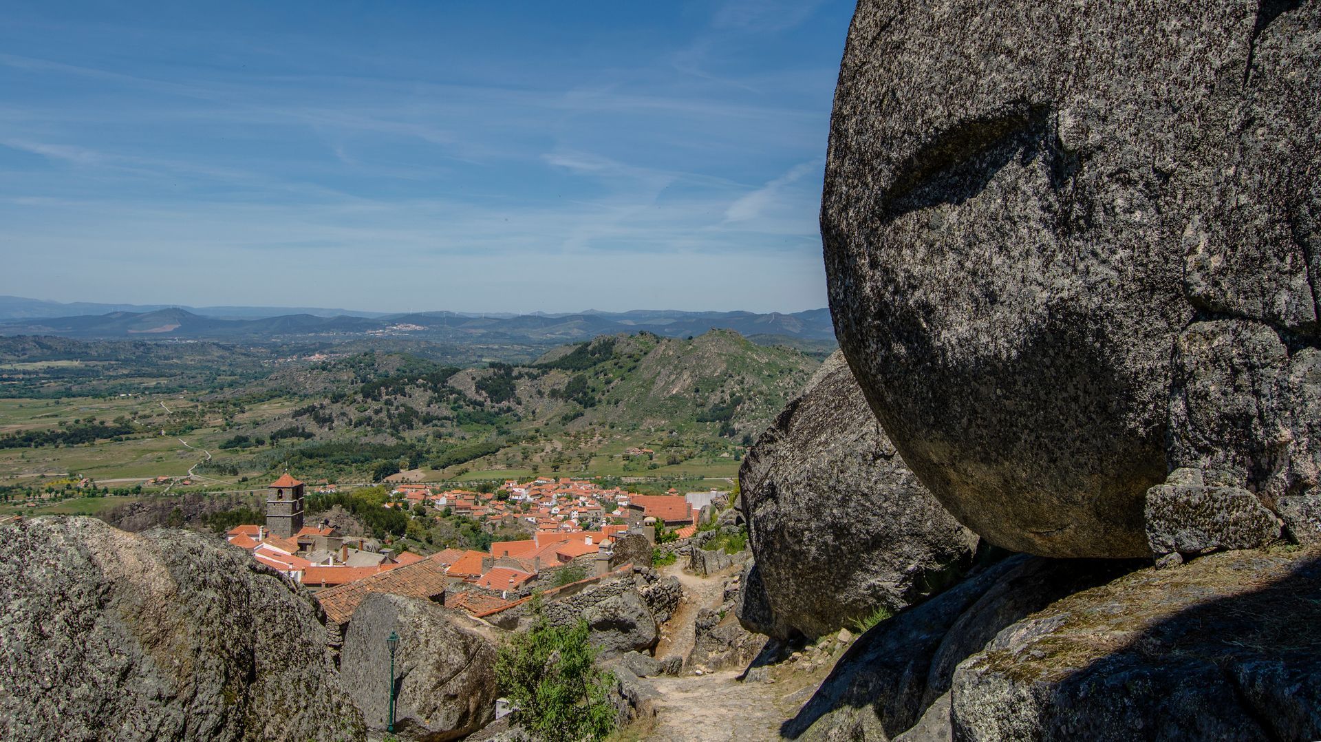 Miradouro rochoso acima de uma aldeia de telhados vermelhos e de um vale verde sob um céu azul