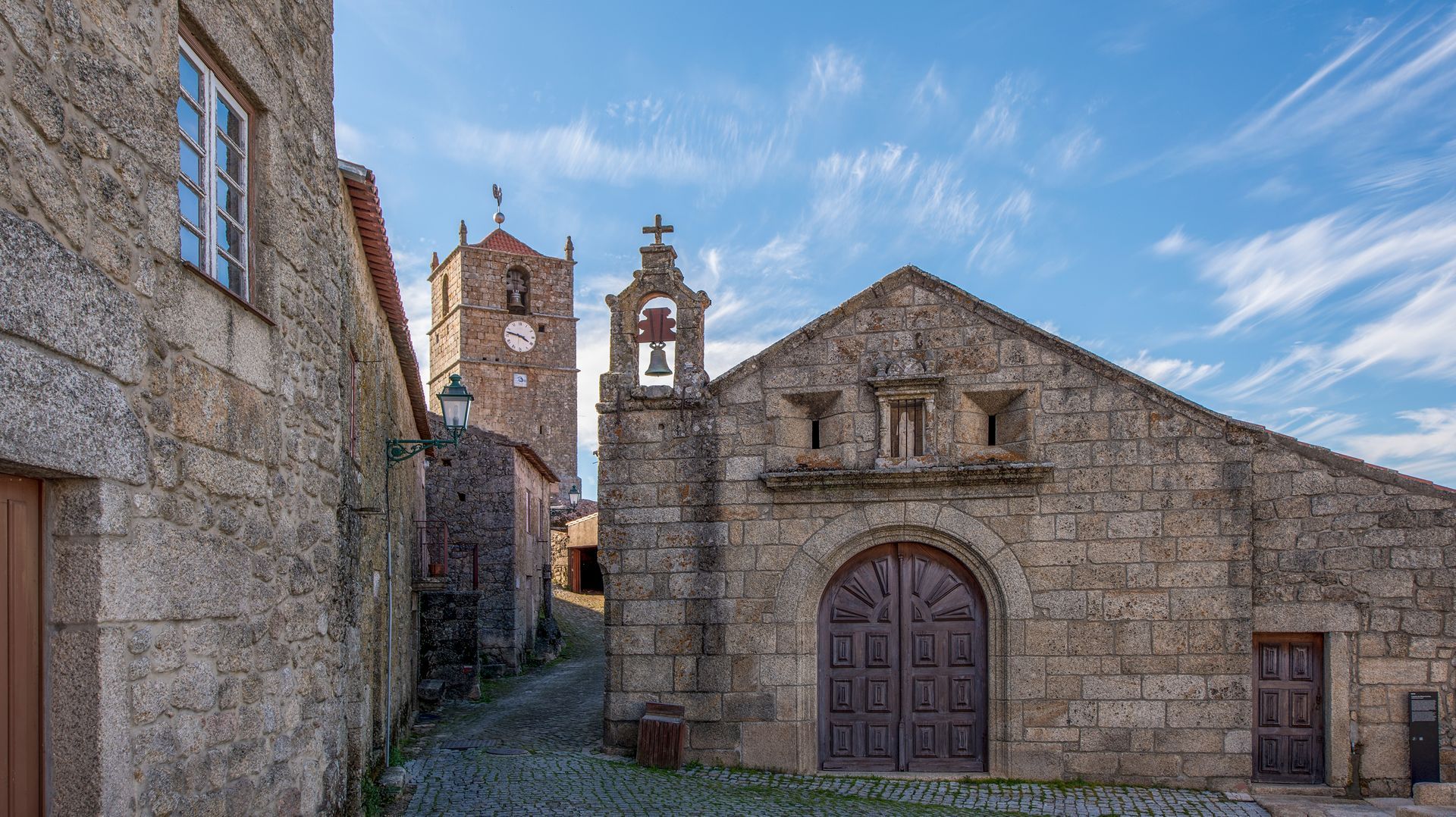 Rua de uma aldeia de pedra, com edifícios rústicos e uma torre sineira sob um céu azul
