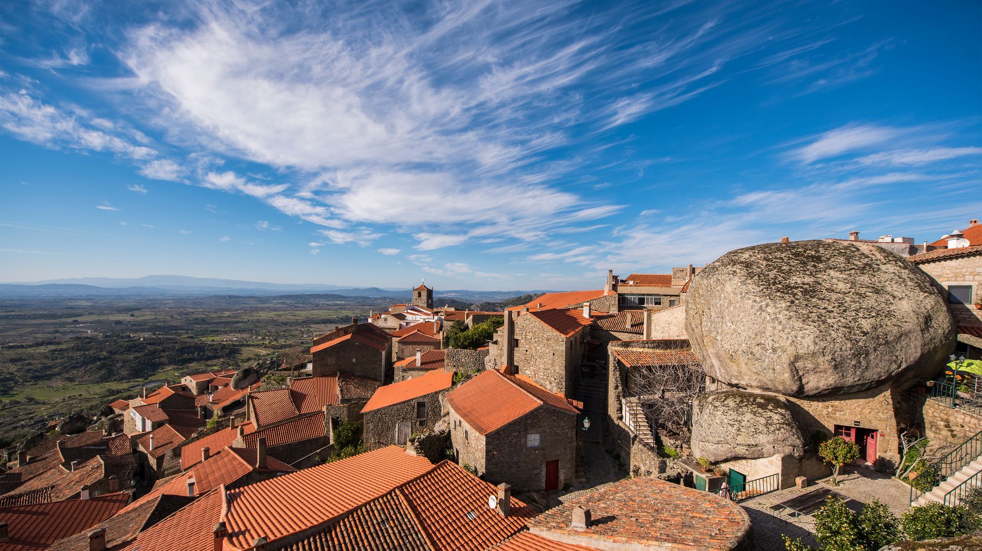 Telhados de pedra de uma aldeia e um grande rochedo com vista para um amplo vale sob um céu azul