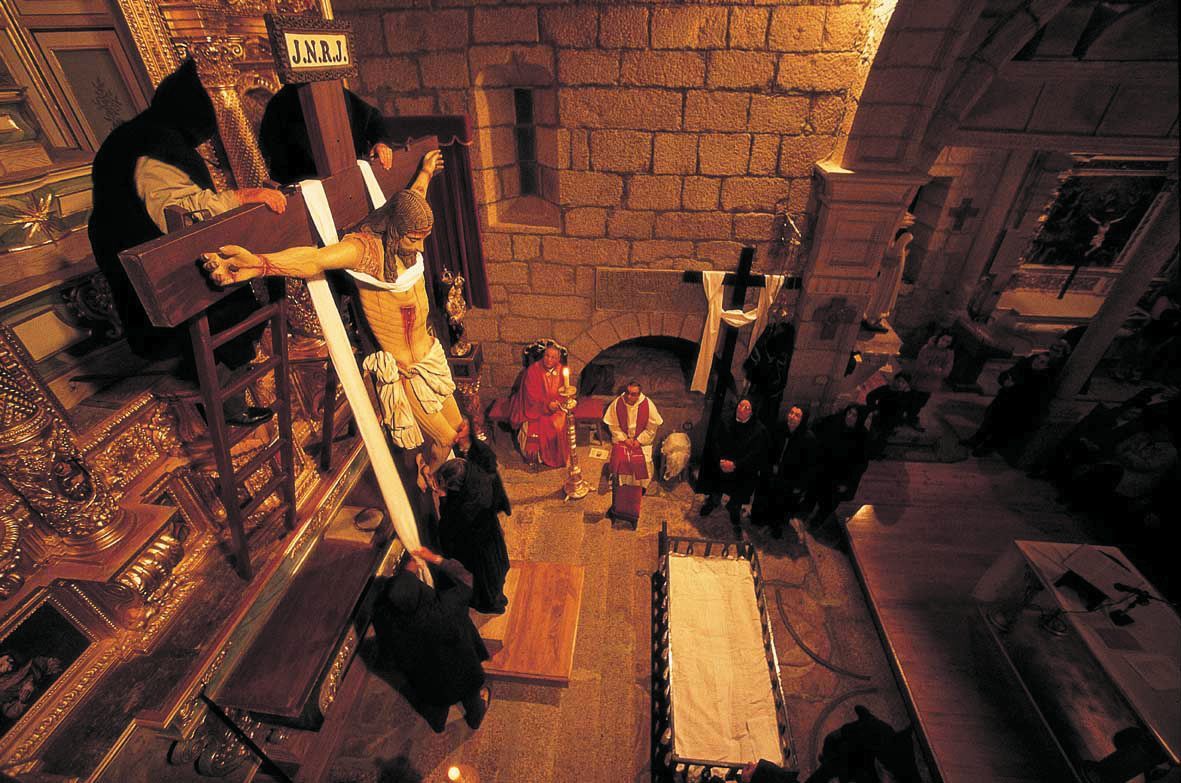 Pessoas vestidas com túnicas escuras carregando grandes cruzes num corredor mal iluminado de uma igreja de pedra