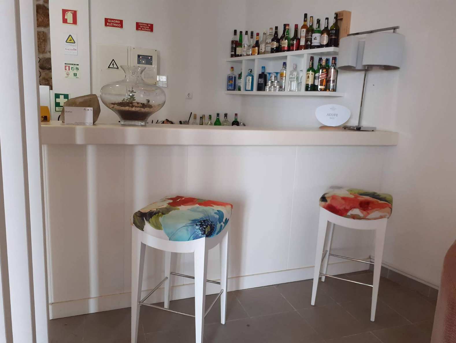 Pequena área de bar em casa, com prateleiras de garrafas, um balcão branco e dois bancos com assentos coloridos.
