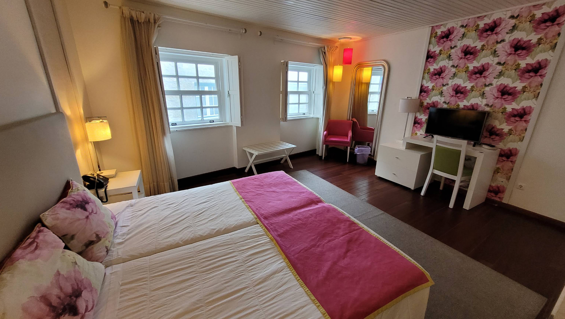 Quarto claro com colcha rosa, papel de parede floral, escrivaninha branca e janelas por onde entra a luz do dia