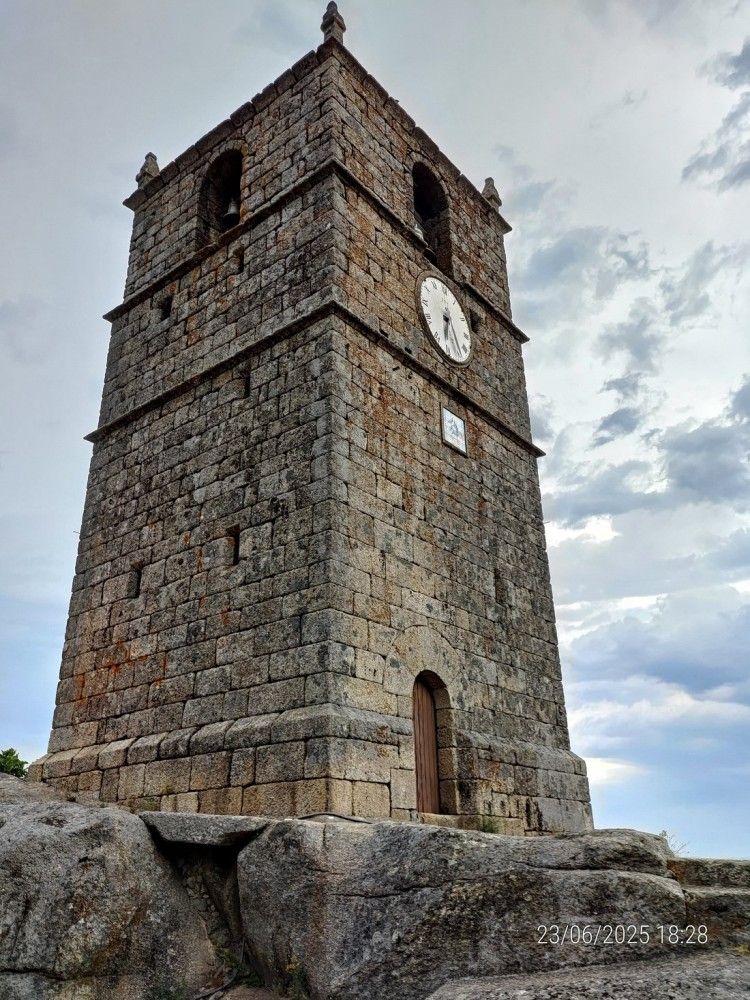 Torre do relógio de pedra no alto de uma colina rochosa, sob um céu nublado