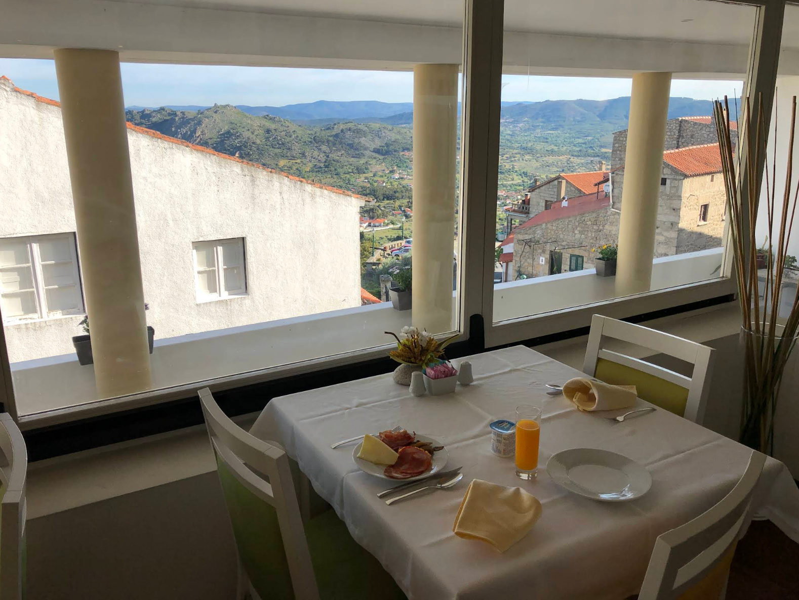 Mesa de jantar numa varanda com vista para uma aldeia ensolarada na encosta e montanhas ao longe
