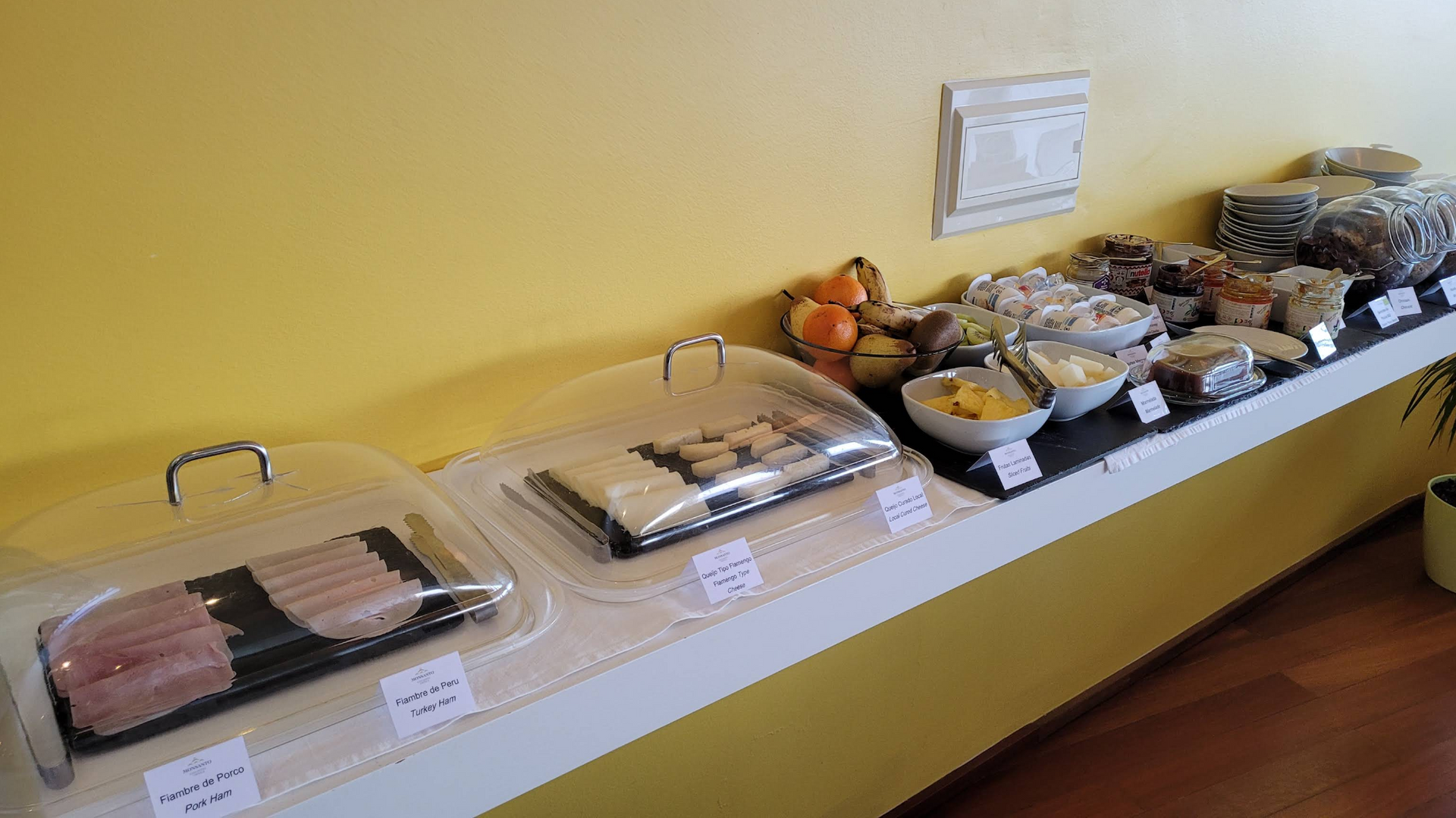 Mesa de bufê com travessas cobertas, frutas, doces e petiscos junto a uma parede amarela