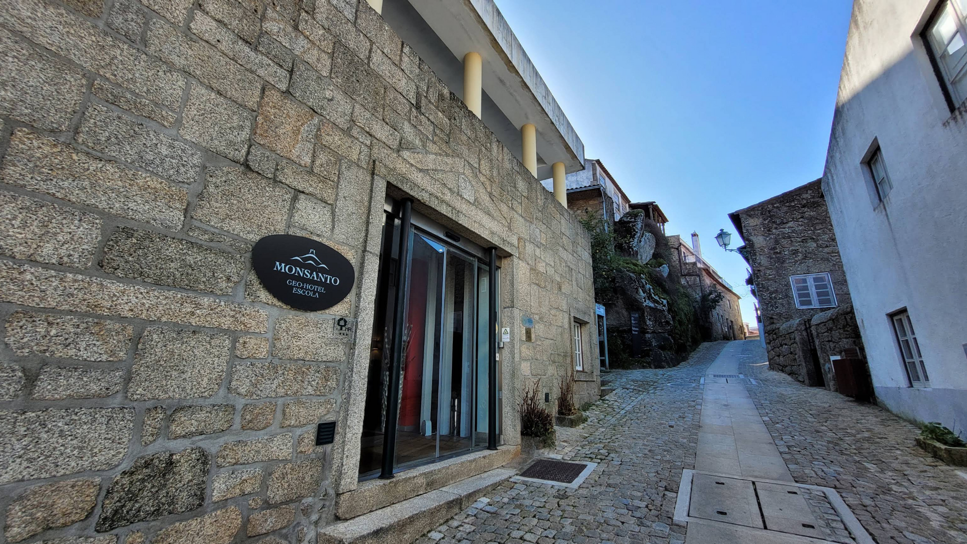 Rua estreita de paralelepípedos entre edifícios de pedra sob um céu azul, com uma loja à esquerda.