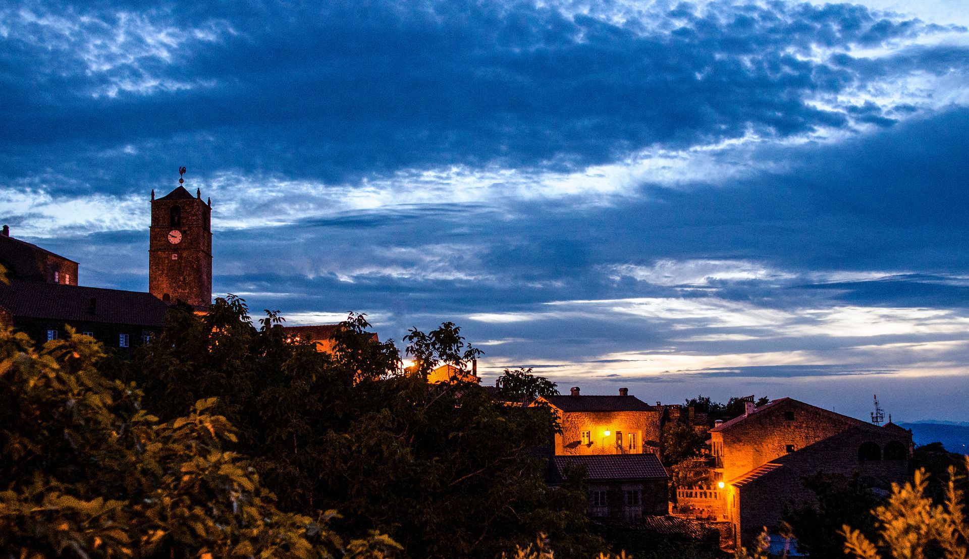 Horizonte ao entardecer sobre uma cidade histórica no alto de uma colina, com luzes e uma torre de pedra.
