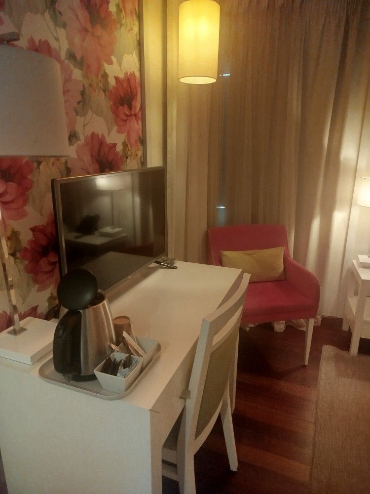 Escrivaninha aconchegante no quarto, com TV, luminária, cafeteira e cadeira rosa ao lado de cortinas translúcidas