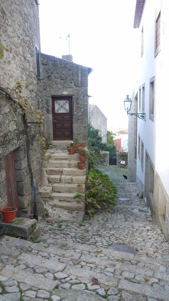 Beco estreito de paralelepípedos entre edifícios de pedra e brancos, com degraus que levam a uma porta de madeira escura.