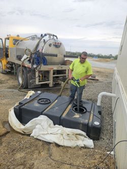 Man in yellow shirt pumping liquid from a black tank into a truck. Outdoors on a construction site.