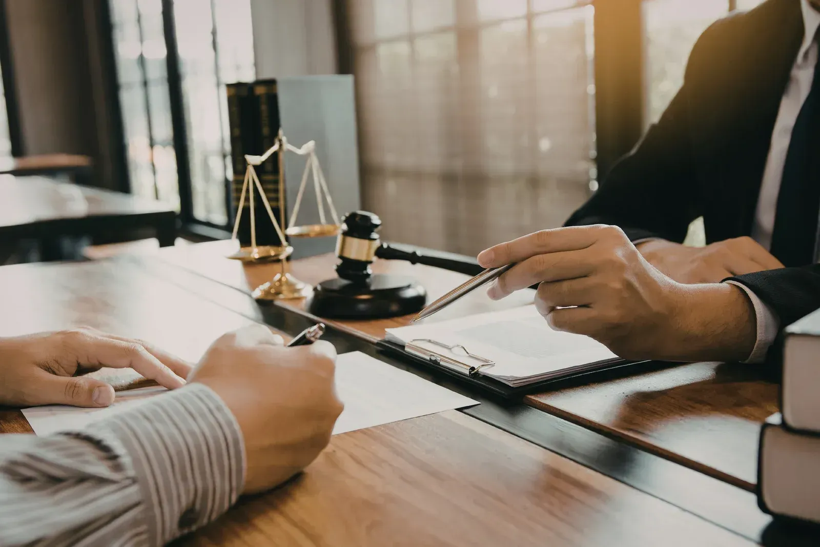 Two people at a desk review documents. One points to a paper. Scales of justice, gavel, books in background.