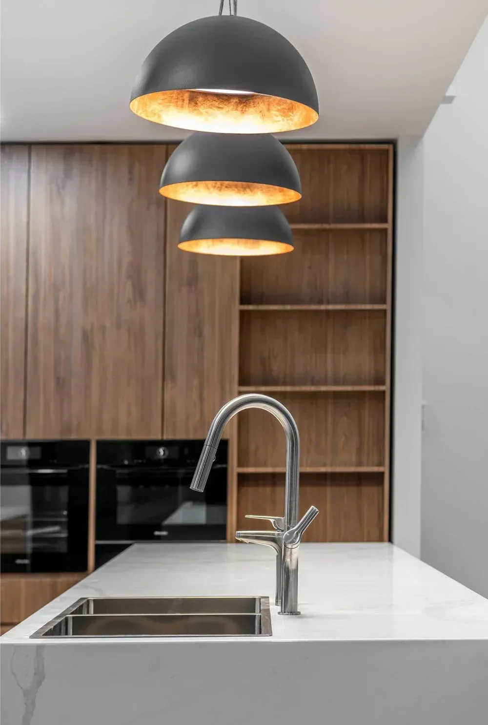 A modern kitchen island with a chrome faucet and sink, illuminated by three stacked dark dome pendant lights.