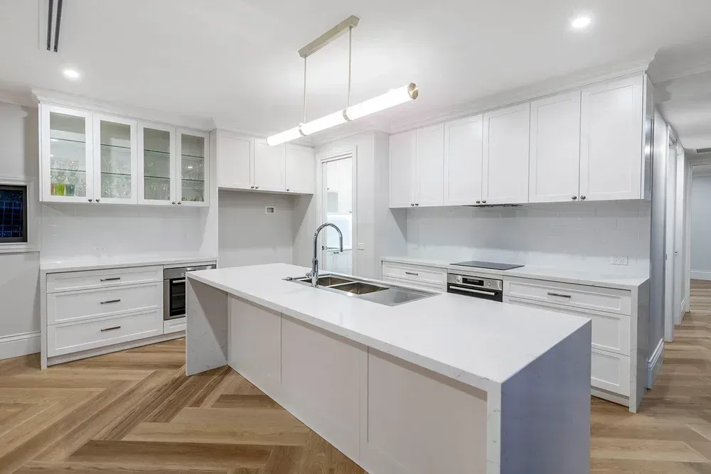 Modern white kitchen with a large island, stainless steel sink, herringbone wood floors, and bright overhead lighting.