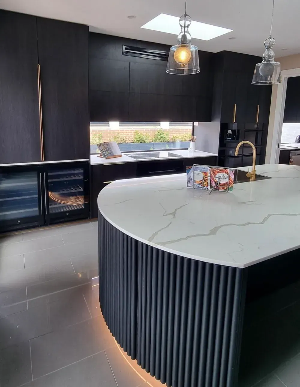A modern kitchen featuring a curved island with dark fluted panels, a white marble countertop, and sleek black cabinetry.