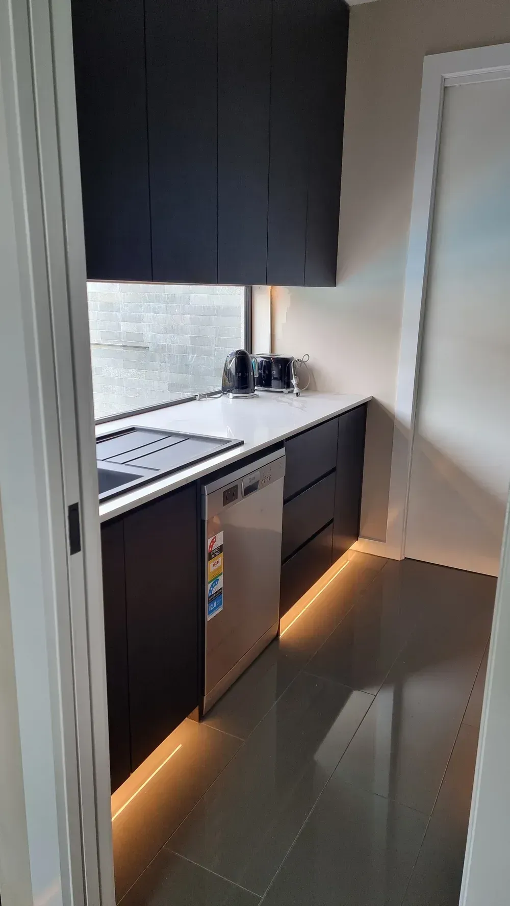 A narrow kitchen galley with dark cabinets, a stainless steel dishwasher, white counters, and under-cabinet lighting.