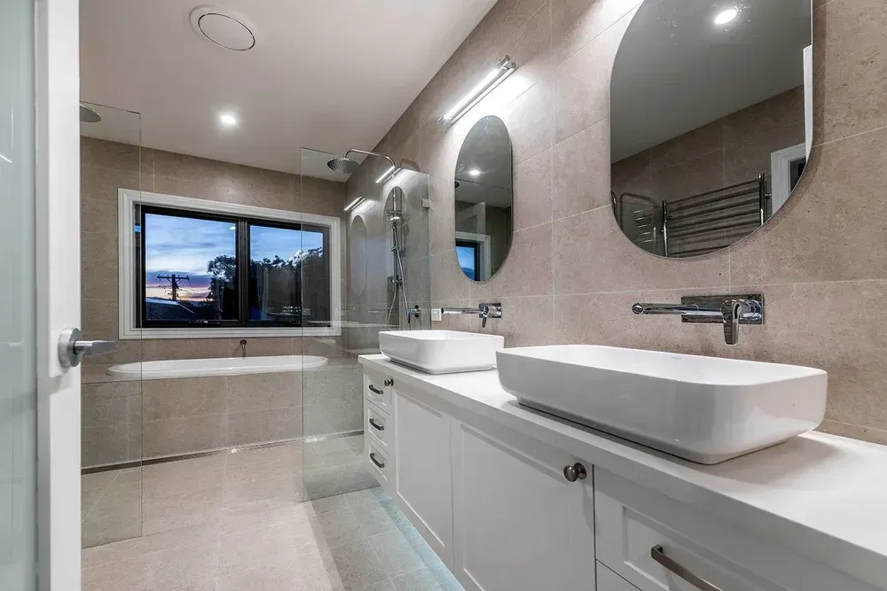 A modern, beige-tiled bathroom featuring a double vanity with white sinks, oval mirrors, and a glass-enclosed bathtub area.