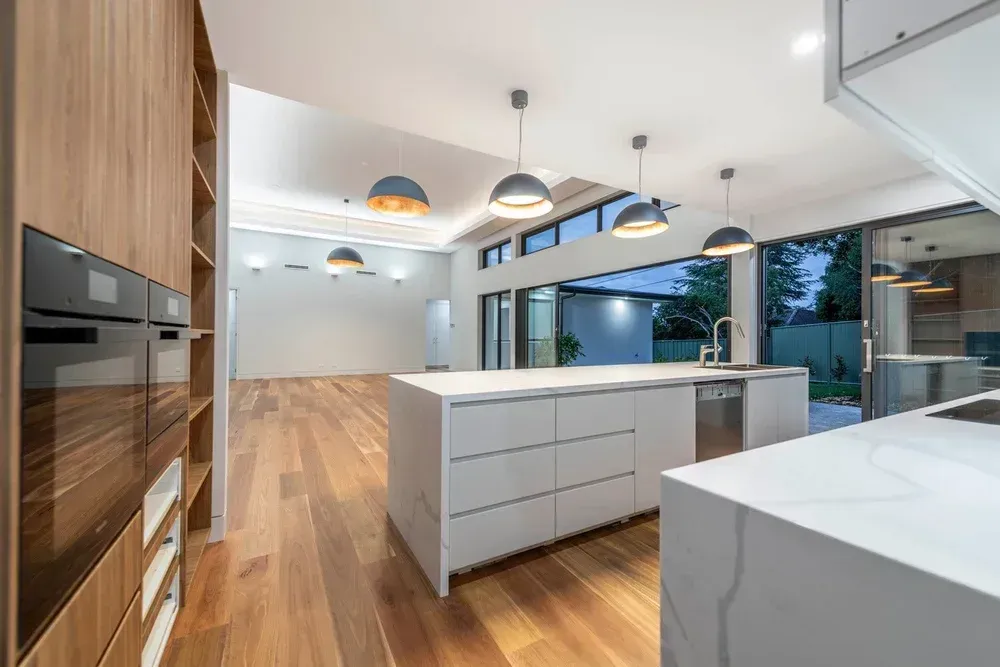 Modern white kitchen with wood floors, island seating, built-in oven, and large windows overlooking a patio