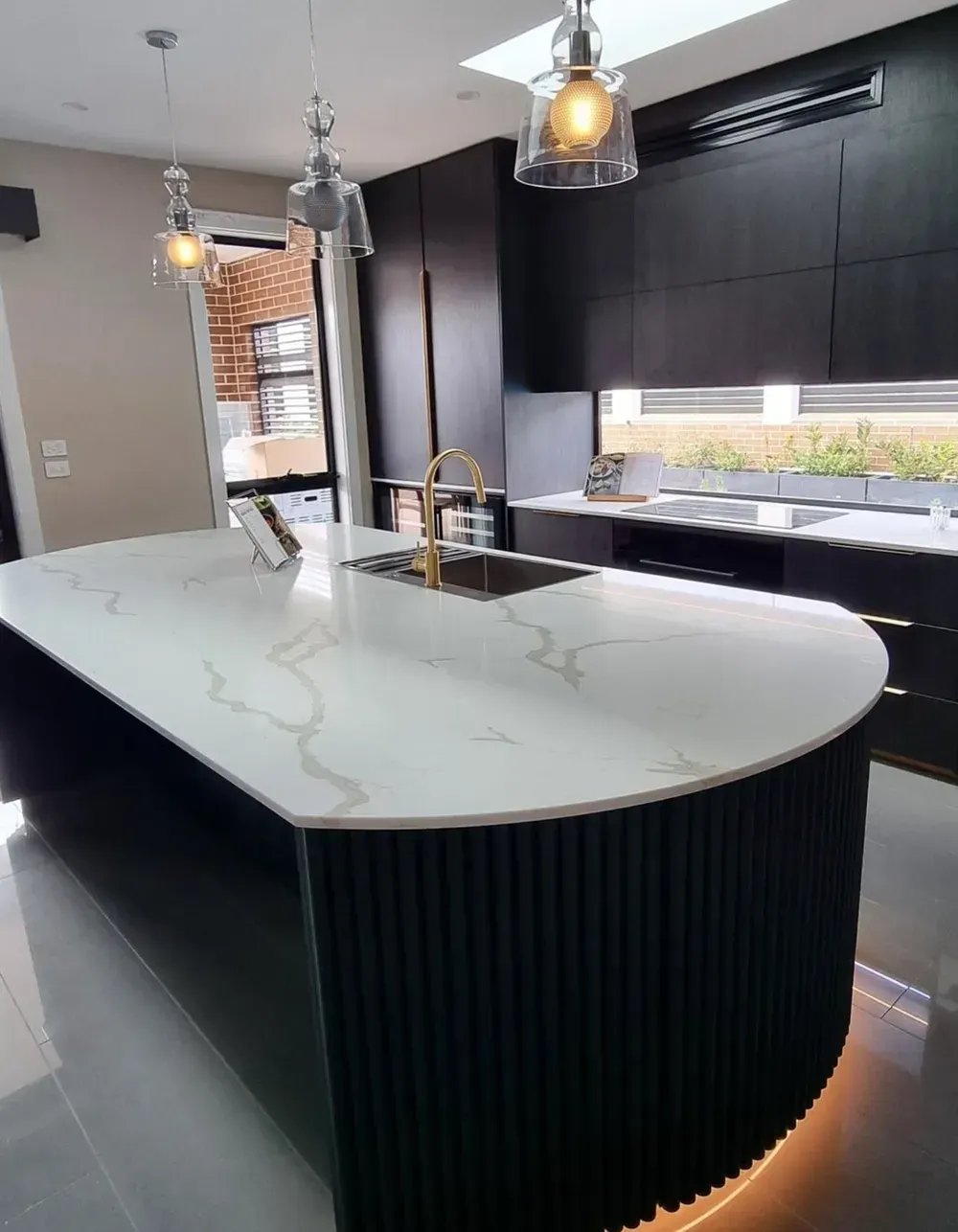 Modern kitchen island with white veined marble countertop, gold sink faucet, and black fluted base with under-lighting.