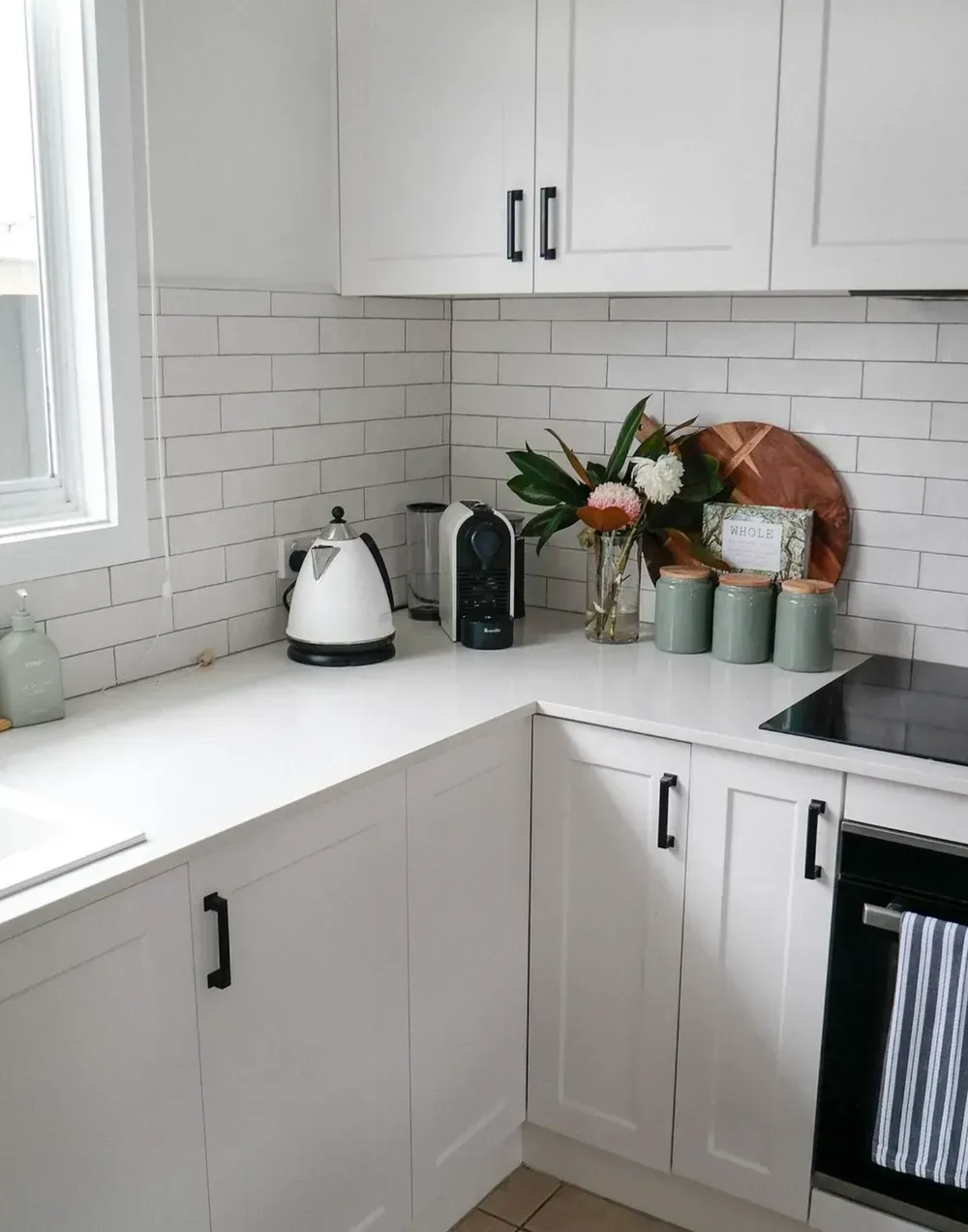 A kitchen corner featuring white cabinets, a white brick-style backsplash, a white kettle, and a small floral arrangement.