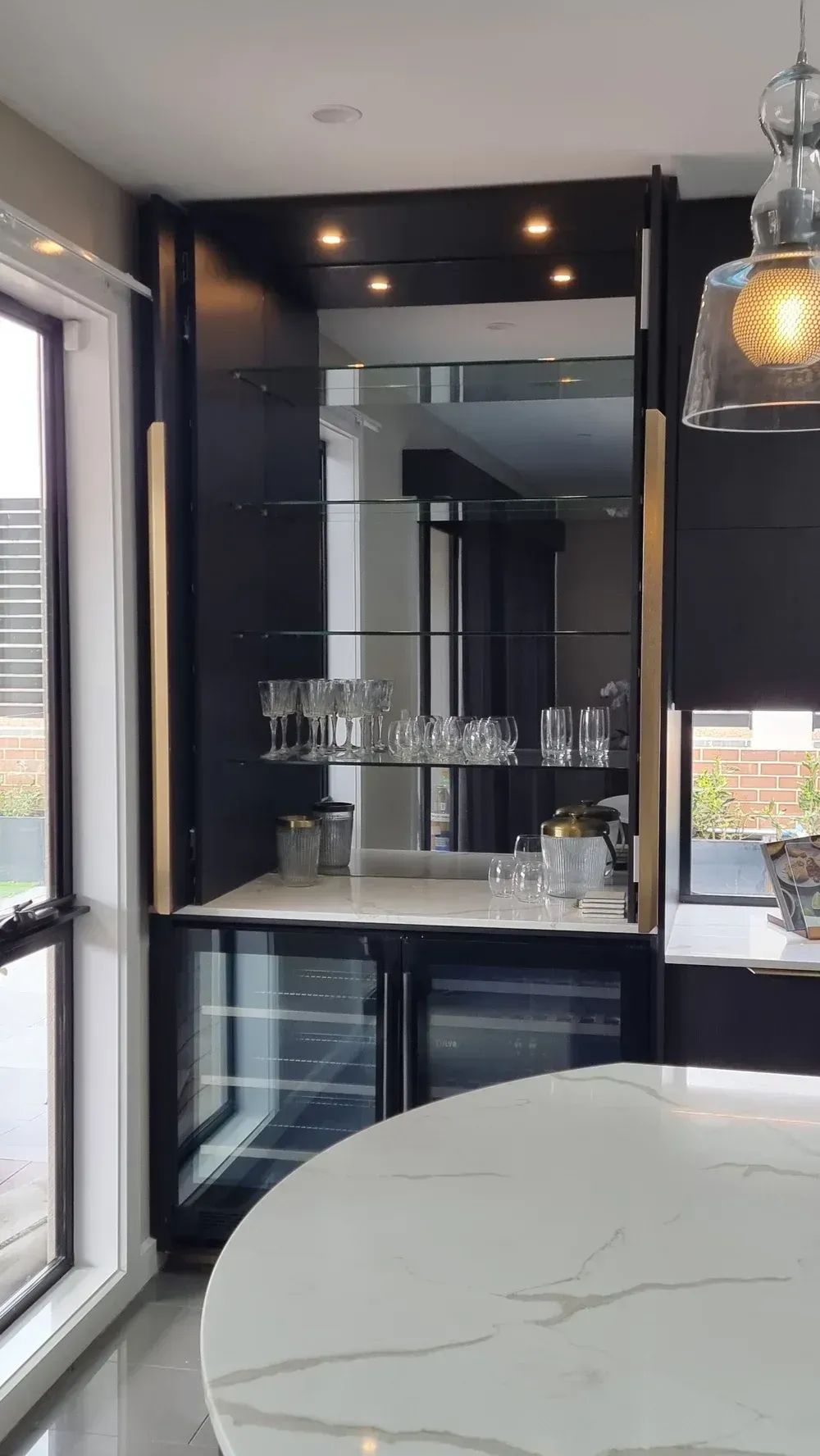 A modern built-in bar with glass shelving, mirror backing, gold accents, and a beverage cooler in a kitchen.