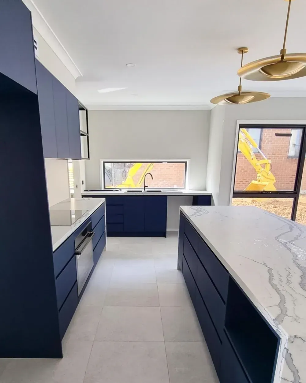 A modern kitchen featuring navy blue cabinets, a marble-topped island, light stone flooring, and two gold pendant lights.