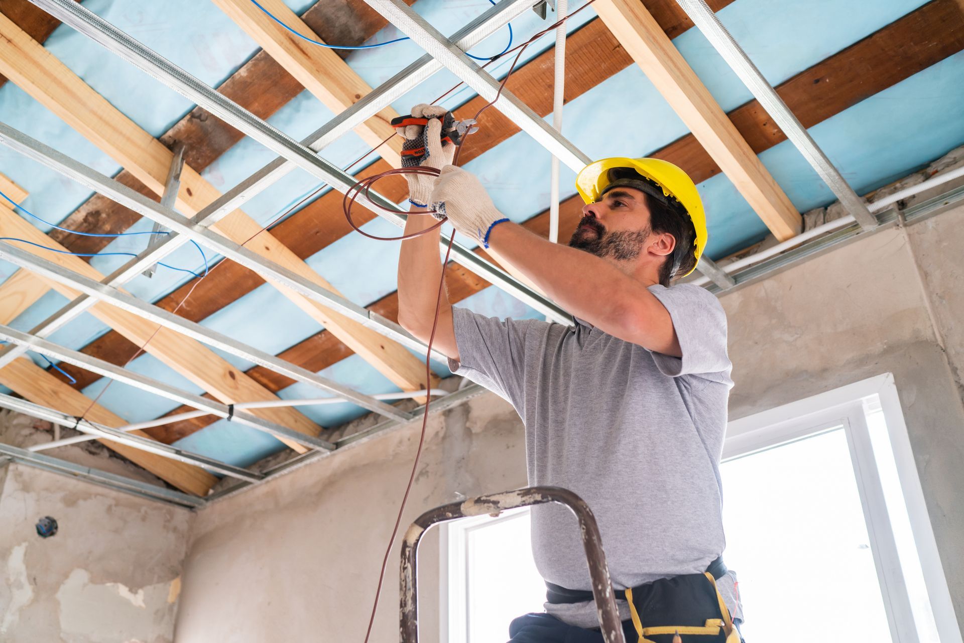 Electrician in yellow hard hat wiring ceiling wires on a ladder inside a building.