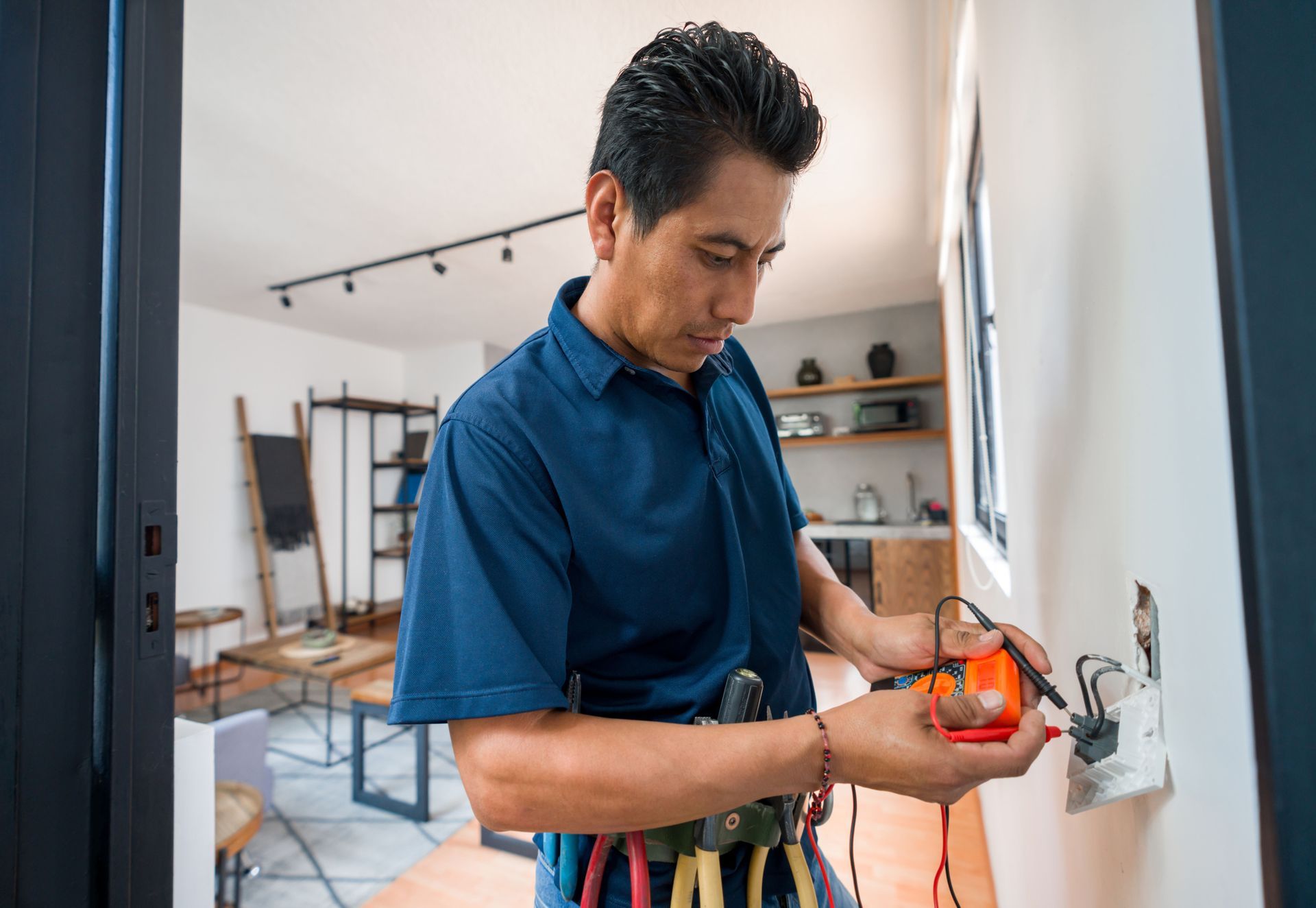 Electrician testing an electrical outlet in a bright room. He's wearing a tool belt and using a meter.