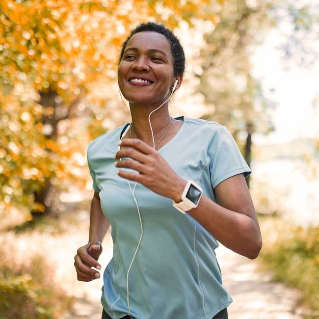 Smiling runner jogging outdoors on a sunlit path, wearing earbuds and a smartwatch.
