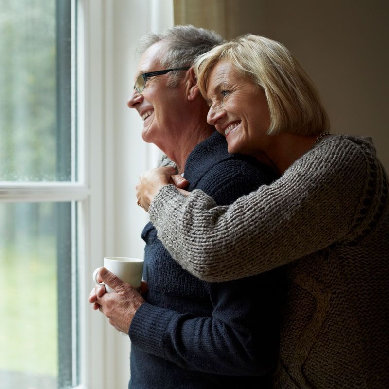 Smiling couple embracing by a window, holding mugs in a cozy room