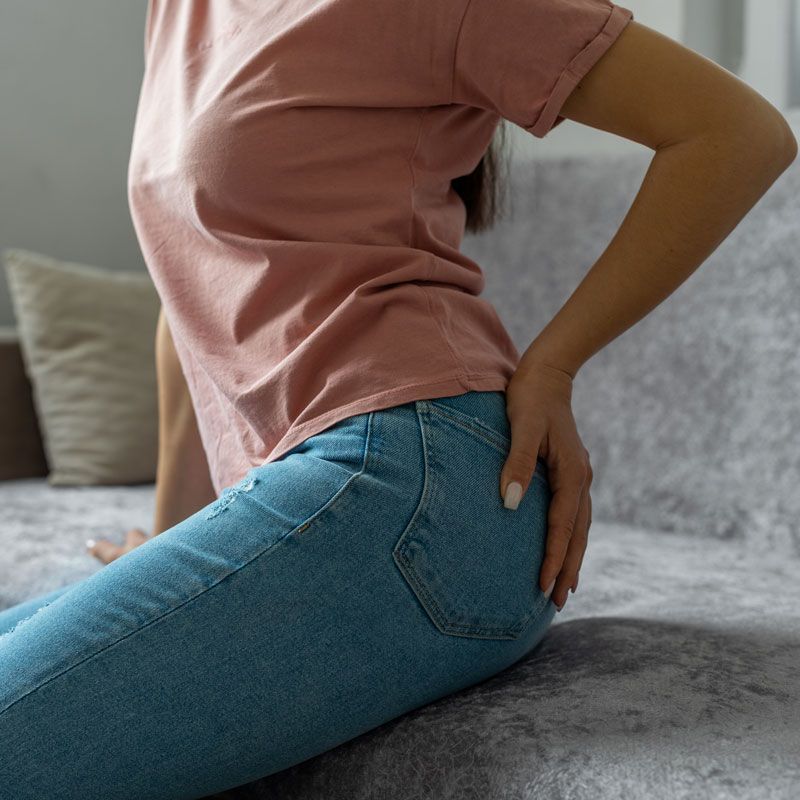 Person in pink shirt and blue jeans holding their lower back while sitting on a bed