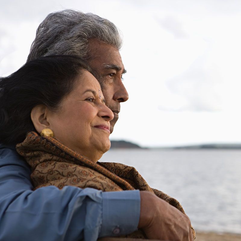 Two people embracing by a calm waterfront, smiling and facing the horizon
