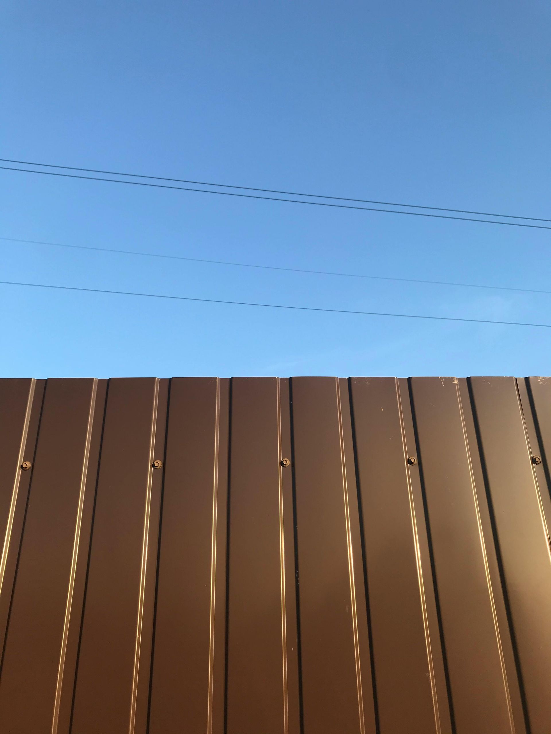 Blue sky above a brown wooden fence with two thin power lines overhead