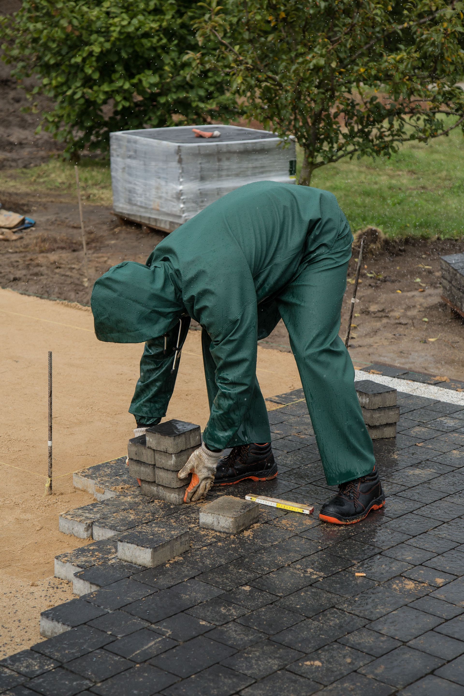 Worker in green overalls laying bricks on a paved path beside a garden.