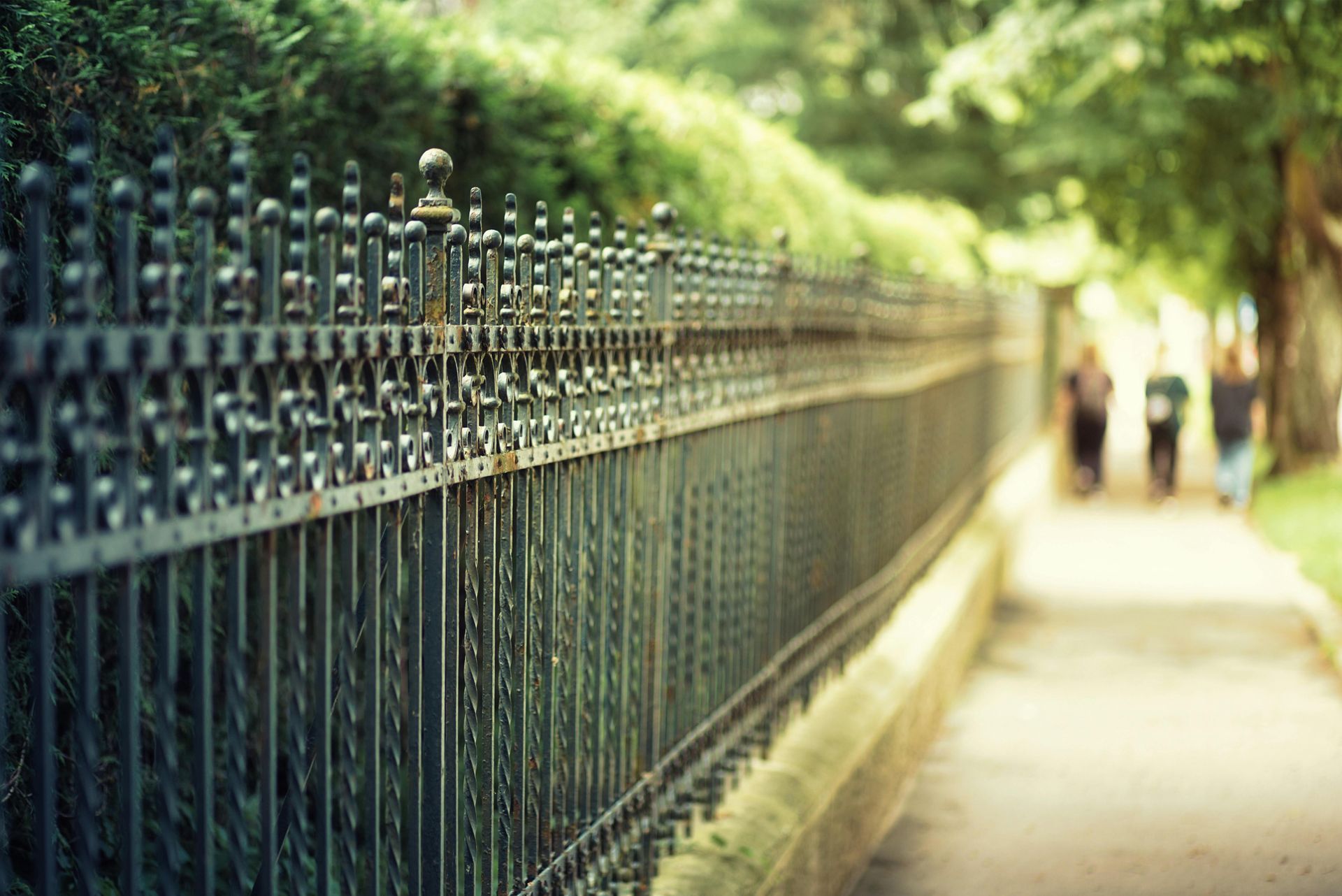 Long black iron fence beside a sunlit path with blurred trees and people in the distance
