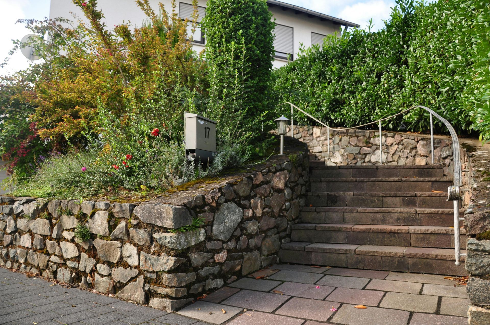 Stone steps beside a lush garden and stone wall leading up to a house with a metal handrail